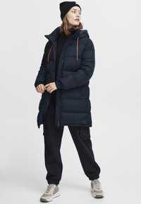 Navy puffer coat with a hood, featuring orange drawstrings, zippered pockets, and ribbed cuffs, paired with black sweatpants and sneakers.