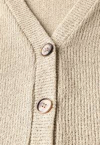 Beige gestrickte Strickjacke mit V-Ausschnitt, ausgestattet mit zwei großen, gemusterten Knöpfen und einem strukturierten Stoff. Nahaufnahmen heben die Maschenmuster hervor.
