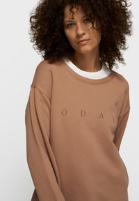 Sweat-shirt marron avec un col blanc, arborant le texte "O D A Y" en imprimé foncé. Tissu lisse et décontracté avec une coupe ample et des manches longues.