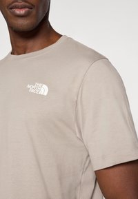 Μπεζ βαμβακερό t-shirt με κοντά μανίκια και στρογγυλή λαιμόκοψη, με λευκό λογότυπο της The North Face στο αριστερό άνω μέρος του στήθους.