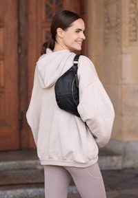 Borsa crossbody nera in pelle con chiusura a zip, texture liscia e tracolla regolabile, abbinata a una felpa oversize beige e leggings chiari.