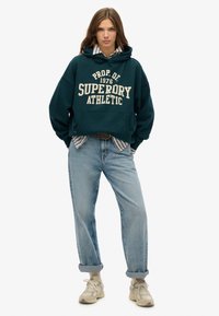 Sudadera verde con el texto "SUPERDRY ATHLETIC", combinada con jeans de manga arremangada en azul claro y zapatillas beige. Camisa blanca de rayas debajo.