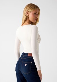 Weißes Langarmshirt mit rundem Ausschnitt und geraffter Vorderseite, kombiniert mit dunkelblauen Jeans mit braunem Lederpatch und einem dreieckigen Logo.