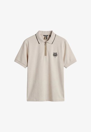 Polo de manga corta beige con cuello ribeteado en negro, cremallera frontal y pequeño logo de cabeza de tigre negra en el pecho izquierdo.