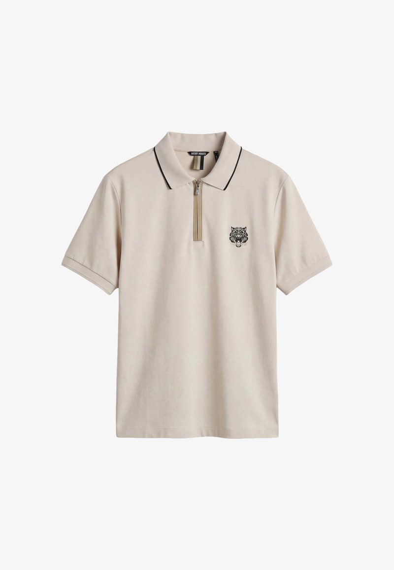 Polo de manga corta beige con cuello ribeteado en negro, cremallera frontal y pequeño logo de cabeza de tigre negra en el pecho izquierdo.