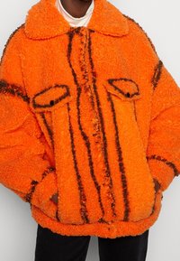 Oranžová huňatá bunda z texturované látky, s límcem, dvěma předními kapsami a černými šikmými pruhy jako akcent.