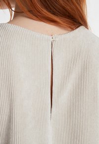 Hellbeige geripptes Oberteil mit strukturierter Oberfläche, das einen vertikalen Schlitz auf der Rückseite und einen einzelnen weißen Knopfverschluss aufweist.