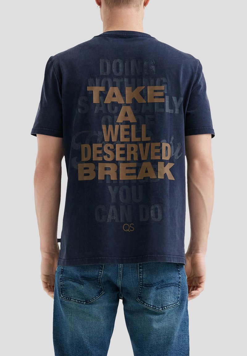 Man in een marineblauwe T-shirt met de tekst "NEEM Een WELL-DESERVED PAUZE" op de achterkant, in combinatie met blauwe jeans, staand tegen een lichte achtergrond.