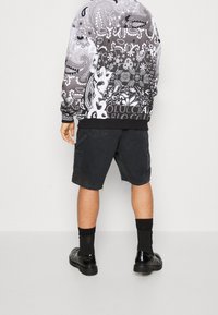 Černobílá mikina s komplikovanými vzory, kombinovaná s tmavými kapsáčovými kraťasy a černými koženými botami, pohled zezadu.