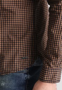 Camisa de cuadros marrón y azul marino con mangas largas, con dobladillo curvado y una etiqueta de marca. El tejido tiene una textura suave. Se lleva con vaqueros azul oscuro.