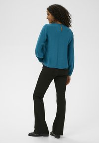 Teal blouse met lange mouwen en knopen detail op de achterkant, gecombineerd met zwarte flared broek en zwarte schoenen, tegen een neutrale achtergrond.