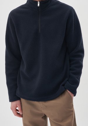 Homme portant un pull en polaire bleu marine avec une fermeture éclair au quart et un pantalon beige, debout une main dans la poche devant un fond uni.