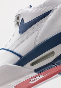 Witte leren sneaker met marineblauwe accenten, voorzien van een opvallende swoosh, textuurdetails en een zichtbare luchtkussenunit in de zool.