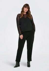 Jauna moteris su ilgais plaukais, vilkinti visiškai juodą aprangą: skaidrią ilgomis rankovėmis palaidinę, juodas kelnes ir aulinukus, stovi su rankomis kišenėse.