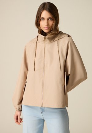 Kobieta o jasnobrązowych włosach, ubrana w beżową kurtkę z kapturem na zamek i jasnoniebieskie dżinsy, stojąca na tle jednolitego tła.