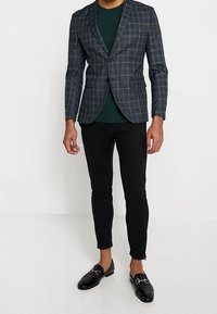 Blazer im dunkelblauen Karomuster mit dünnen weißen Linien, schwarze enganliegende Hose und glänzende schwarze Slipper. Darunter ein grünes T-Shirt.