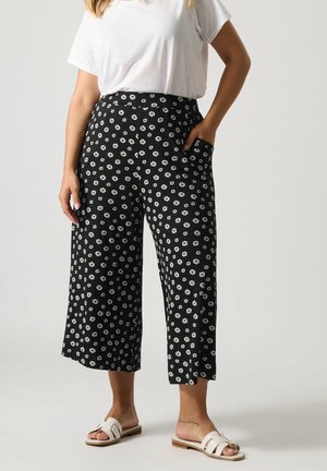 Kobieta ubrana w czarne spodnie o szerokich nogawkach z białym nadrukiem stokrotek, białą koszulę z krótkim rękawem oraz białe klapki, stojąca z jedną ręką w kieszeni.