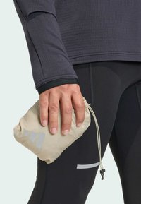 Béžová taška so sťahovacou šnúrkou, držaná v ruke. Osoba má na sebe tmavo dlhý rukáv a čierne športové nohavice s reflexnými akcentmi.