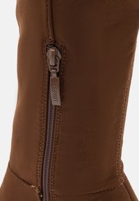 Botín de ante marrón hasta el tobillo con cremallera lateral, que presenta un acabado texturizado y un tacón bajo. El herraje tiene un logo en relieve para mayor detalle.