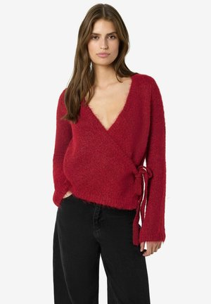 Woman wearing a deep red fuzzy wrap sweater tied at the side paired with loose black pants against a plain white background.