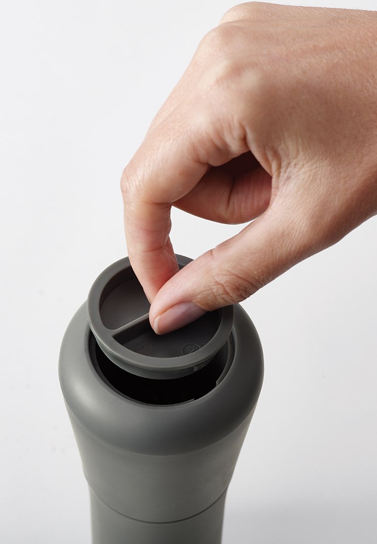 Een hand tilt een ronde grijze deksel van de bovenkant van een bijpassende grijze cilindrische container tegen een eenvoudige witte achtergrond.