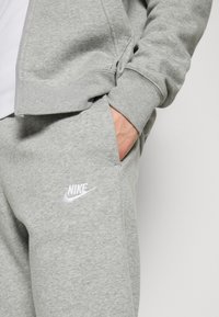 Šedé Nike tepláky mají měkkou, strukturovanou tkaninu s elastickým pasem a bílým logem na levém stehně.
