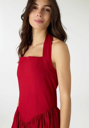 Donna con lunghi capelli scuri che indossa un vestito rosso senza maniche con scollo quadrato, in piedi davanti a uno sfondo bianco semplice.