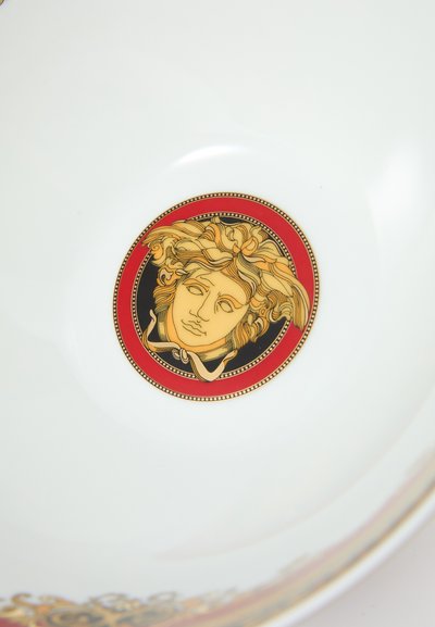 Λευκό κεραμικό μπολ με κεντρικό κυκλικό σχέδιο ενός προσώπου σε χρυσούς τόνους, περιβαλλόμενο από κόκκινο και μαύρο περίγραμμα με διακοσμητικές λεπτομέρειες.