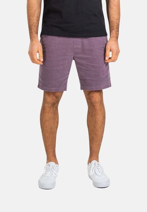 Unterkörper einer Person, die lila Shorts, ein schwarzes Hemd und weiße Turnschuhe trägt, steht mit entspannt an den Seiten hängenden Händen vor einem einfarbigen Hintergrund.