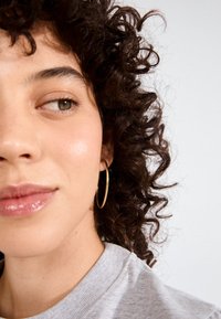 Tuvplāns ar seju ar cirtainiem matiem, valkājot lielas zelta auskariņu gredzenus, gaiši pelēku džemperi un spīdīgas lūpas. Gluda ādas tekstūra, neitrāla fona.