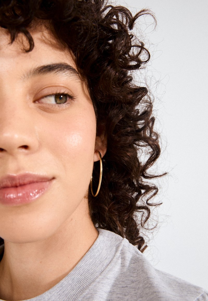 Tuvplāns ar seju ar cirtainiem matiem, valkājot lielas zelta auskariņu gredzenus, gaiši pelēku džemperi un spīdīgas lūpas. Gluda ādas tekstūra, neitrāla fona.