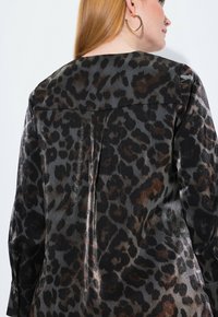Blusa con estampado de leopardo y una textura satinada suave; cuenta con un cuello clásico y un plisado en la parte posterior, en tonos de negro y marrón.