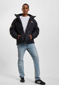 Doudoune noire rembourrée avec fermeture éclair, grande capuche et logo sur la poitrine ; portée sur un t-shirt blanc et assortie à un jean bleu clair.