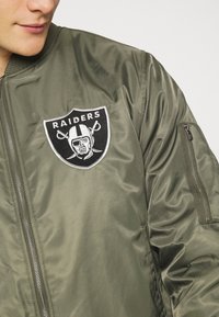 Olive green bomber jacket featuring a black and white embroidered Raiders logo on the chest and a zippered pocket on the left sleeve.