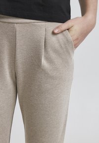 Pantalon beige texturé avec des plis à l'avant et des poches latérales, associé à une chemise noire. Le tissu semble doux et lisse.