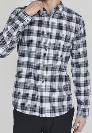 Hombre ajustando el cuello de una camisa de cuadros en negro, blanco y gris, usando pantalones negros, con fondo gris liso.