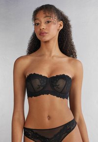 Soutien-gorge sans bretelles noir avec des détails en dentelle et des bonnets en tulle, présentant un bord festonné. Sous-vêtements en dentelle noire complémentaires visibles en dessous.
