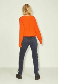 Jasnopomarańczowy sweter ribbed, luźny krój, w połączeniu z ciemnoniebieskimi dżinsami skinny. Tło jest jasne i neutralne. Widać czarne, masywne buty.