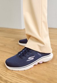Navy blue Skechers slip-on shoes with mesh upper, padded collar, and white sole featuring purple accents, set against light-colored trousers.