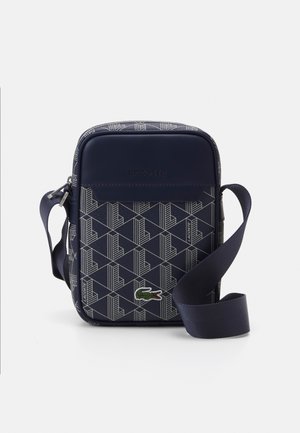 Bolso de hombro azul marino con patrón geométrico, material de cuero elegante, correa ajustable y acento del logo de Lacoste en la parte frontal.