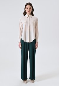Blusa rosa chiaro con due tasche sul petto, colletto e maniche lunghe. Abbinata a pantaloni a gamba larga verde scuro e scarpe beige a punta.