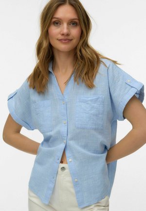 Femme aux cheveux châtain clair mi-longs portant une chemise bleu clair à manches courtes boutonnée et un pantalon de couleur claire, debout les mains sur les hanches.