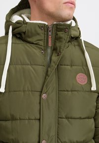 Olivgrüne Steppjacke mit gefütterter Kapuze, Druckknöpfen und einem strukturierten Patch-Logo auf der Brust. Ausgestattet mit Kordelzügen und einem Reißverschluss.