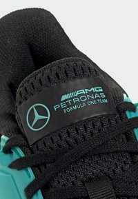 Chaussure de sport noire avec une tige en maillage texturé, présentant un accent turquoise et une étiquette noire avec un texte blanc et le logo Mercedes.
