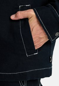 Ciemnoniebieska kurtka z białymi niciami, detalami kieszeni i metalowym guzikiem. Materiał jest teksturowany i trwały.