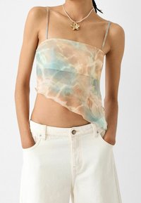 Crop top asimmetrico e trasparente in tonalità pastello di blu e pesca, con spalline sottili e una texture fluida. Abbinato a pantaloni bianchi a vita alta.