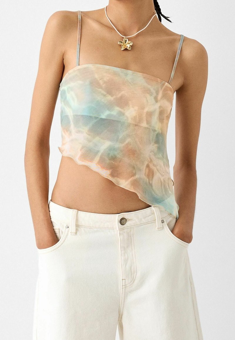 Crop top asimmetrico e trasparente in tonalità pastello di blu e pesca, con spalline sottili e una texture fluida. Abbinato a pantaloni bianchi a vita alta.