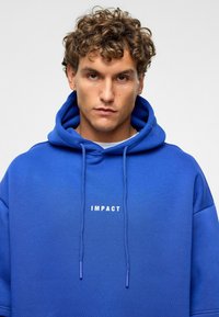 Blauwe hoodie van zachte stof, met een voorzak in kangoeroejasje en aantrekkoorden. "IMPACT" in het wit gedrukt over de borst.
