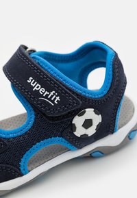 Sandalia azul marino con material de malla, sujeta con una correa de Velcro. Presenta acentos en azul y una decoración de balón de fútbol en el lateral.