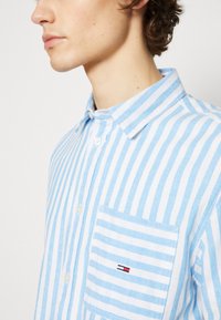 Camicia a righe blu chiaro e bianco, con colletto a punta, taschino frontale con un piccolo logo e un tessuto liscio e strutturato.